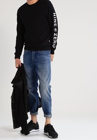 Svart sweatshirt med "NINE ZERO" tryck på ärmarna, i kombination med ljusblå denimjeans och svarta sneakers, håller en svart jacka.