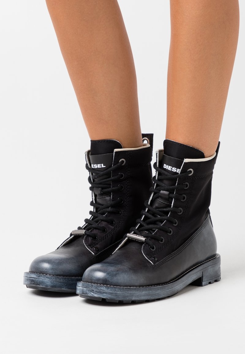 Bottes de combat montantes en cuir noir avec lacets, logo Diesel sur la languette, portées sur des jambes nues sur un fond blanc.