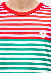 Egy személy nyaka és felső mellkasa, aki piros, fehér és zöld csíkos inget visel, piros gallérral és egy kis fehér virággal a mellkasán.