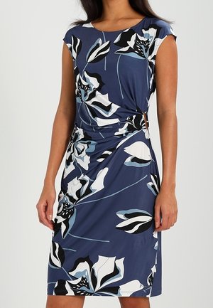 Kobieta ubrana w granatową sukienkę bez rękawów z dużym białym, czarnym i jasnoniebieskim kwiatowym wzorem, z marszczeniem po bokach i o długości do kolan.