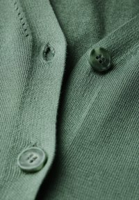 Maglione lavorato a maglia verde con una trama liscia, caratterizzato da due bottoni rotondi di un verde scuro e un motivo di cucitura sottile.