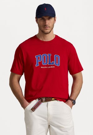 Tricou roșu din bumbac cu logo-ul "POLO" în albastru și alb, asortat cu pantaloni albi și o curea multicolorată în dungi. Căciulă bleumarin pe cap.