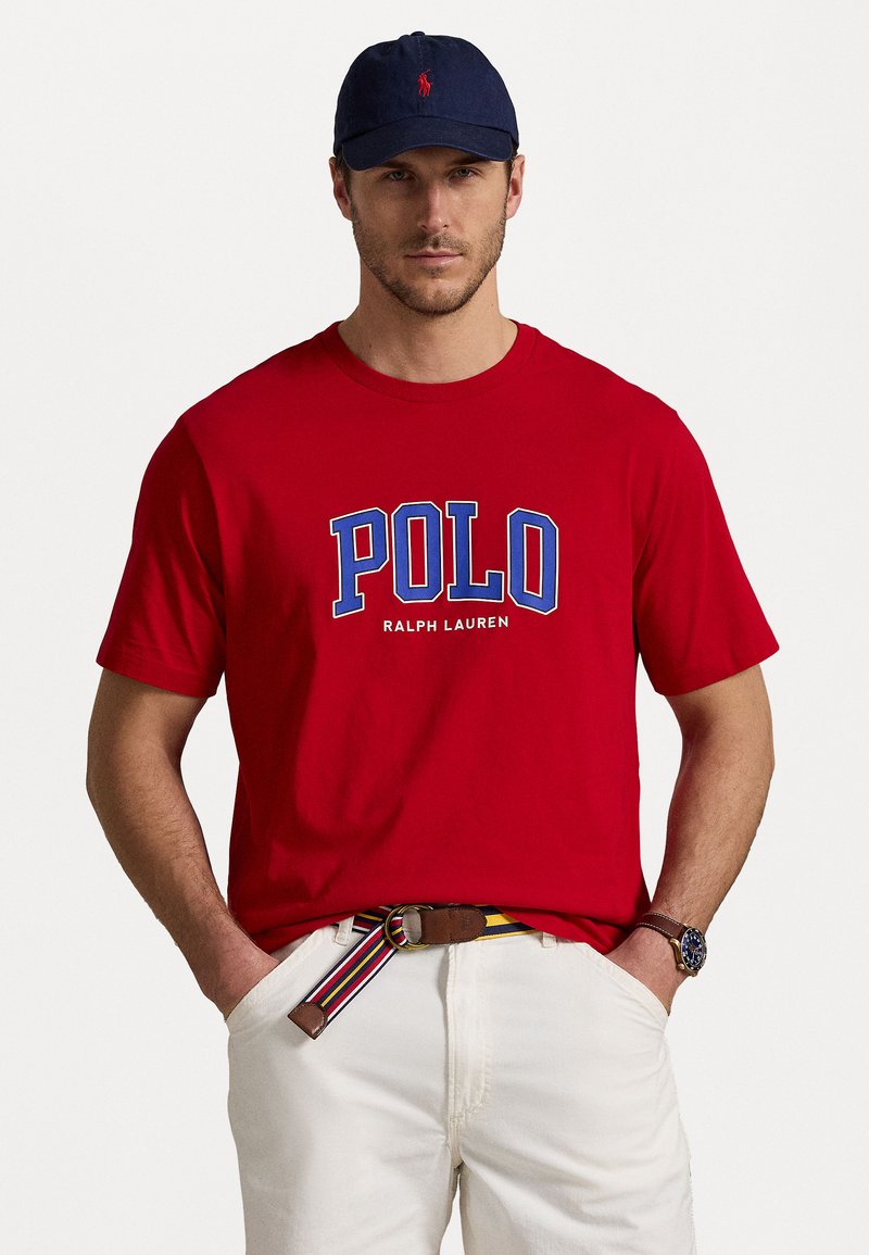 Red cotton t-shirt with "POLO" logo in blue and white, paired with white pants and a multicolored striped belt. Navy cap on head.