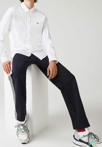 White button-up shirt with long sleeves and a small green logo. Paired with navy pants featuring white stripes and multi-colored sneakers.