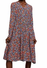 Pitkähiha, V-pääntie mekko, jossa on kellotettu hame tiheällä kukkakuviolla punaisissa, purppuran, valkoisissa ja mustissa väreissä.