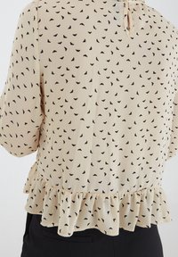 Blusa beige con motivo a foglia nera, tessuto testurizzato, orlo arricciato e chiusura con bottone sul retro, con maniche ampie.