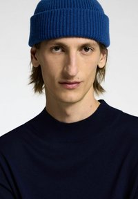 Maglione a coste di colore blu navy con colletto rotondo alto; indossato con un berretto a coste di colore blu brillante. Mostrato il torso superiore di un modello.