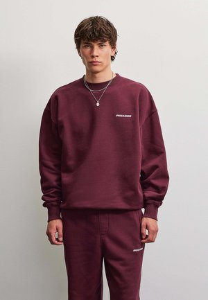 Young man with short curly hair wearing a matching maroon sweatshirt and pants with white "PEGADOR" logos on chest and thigh.