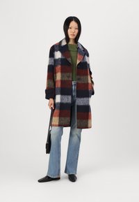 Manteau en laine à carreaux rouge, marine et blanc, avec un large col, deux boutons et de grandes poches. Associé à un jean évasé et des chaussures noires.