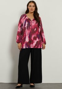 Langärmlige Bluse mit pink-lila Muster, Henley-Ausschnitt und Knopfdetails, kombiniert mit schwarz-weißen weit geschnittenen Hosen und flachen Schuhen.