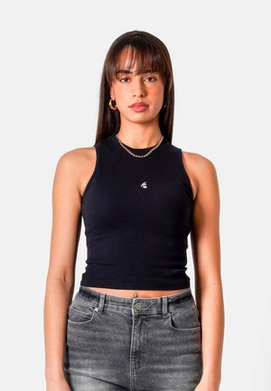 Junge Frau mit braunen Haaren, die Creolen, eine goldene Halskette, ein schwarzes ärmelloses Oberteil und graue Jeans vor schlichtem Hintergrund trägt.