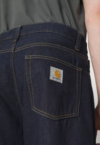 Granatowe dżinsy z żółtymi przeszyciami, z naszywką z logo Carhartt na tylnej kieszeni. Zbliżenie na pasek i kieszeń.