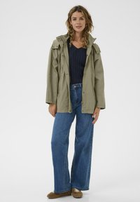 Svetlo olivno zelena jakna s kapuco, z zapenjanjem na gumbe in zadrgo, kombinirana z visokim pasom modrih kavbojk in priložnostnimi rjavimi čevlji.