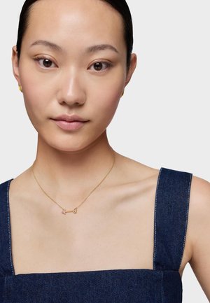 Donna asiatica con pelle liscia che indossa piccoli orecchini d'oro, una collana con ciondolo a freccia d'oro e un top senza maniche in denim blu scuro.