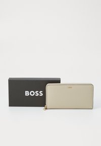 Světle béžová kožená peněženka se zipovým uzávěrem a zlatými detaily. Představena v černé krabičce s logem "BOSS" v bílé barvě.