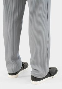 Graue, wellige Hose mit lockerer Passform, kombiniert mit schwarzen Sneakers mit weißen Sohlen und Schnürsenkeln, von hinten gesehen.