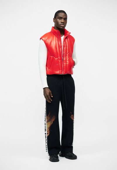 Άνδρας που στέκεται, φοράει κόκκινο αμάνικο φουσκωτό γιλέκο πάνω από λευκό μακρυμάνικο πουκάμισο, μαύρο παντελόνι με φλόγες και λεπτομέρειες κειμένου, και μαύρα αθλητικά παπούτσια.