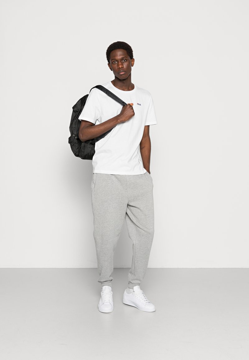 Camiseta blanca de manga corta con logo, pantalones de chándal grises, mochila negra y zapatillas blancas, de pie sobre un fondo liso.