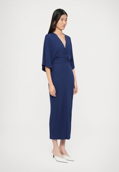 Μπλε σκούρο φόρεμα με βαθύ V-λαιμόκοψη, μανίκια με κυματιστό σχέδιο και ρυτιδώσεις στο μπούστο, συνδυασμένο με λευκές μίνι πλατφόρμες με τακούνι.