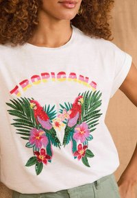 T-shirt en coton blanc avec un motif brodé coloré représentant deux perroquets rouges, des feuilles tropicales et des fleurs, avec "TROPICANA" en lettres dégradées.