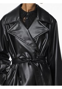 Trench-coat noir en cuir synthétique avec larges revers, boutons croisés et ceinture en tissu. Présente une texture brillante et une coupe ajustée.
