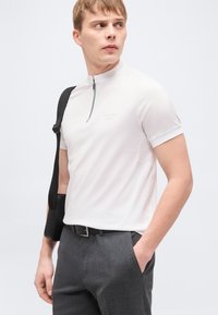 Νεαρός άνδρας που φοράει λευκό κοντομάνικο πουκάμισο με φερμουάρ, γκρι παντελόνι, μαύρη ζώνη και μαύρη τσάντα ώμου, κοιτάζει προς τα αριστερά του.