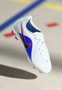 Λευκό ποδοσφαιρικό παπούτσι με μπλε πάνελ και μαύρο-ροζ γραμμές, γωνιασμένο στη μέση του αέρα πάνω από μια ανοιχτόχρωμη επιφάνεια με χρωματιστές γραμμές γηπέδου.