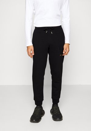 Homme portant un pantalon de jogging noir avec une taille à cordon, des baskets noires et une chemise blanche à manches longues, debout devant un fond uni.