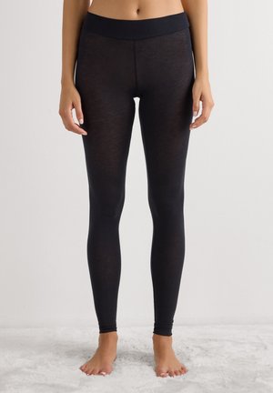 Primer plano de una persona con leggings negros ajustados, de pie descalza sobre una superficie blanca texturizada frente a un fondo claro liso.