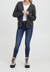 Šedý pletený cardigan s knoflíkovou légou vpředu, doplněný modrými úzkými džíny a béžovými šněrovacími botami. Pod tím jednoduché bílé tričko.