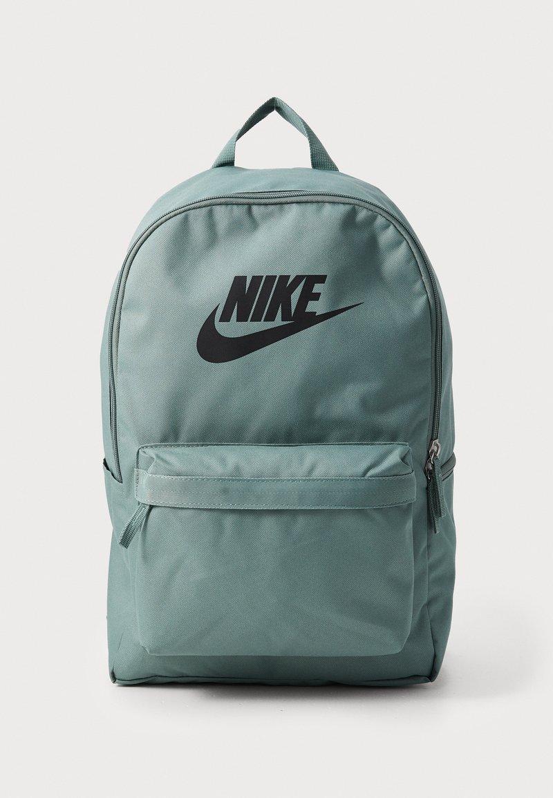 Mochila de color teal claro hecha de tela duradera. Presenta el logotipo negro de Nike, un bolsillo frontal con cremallera y un asa superior acolchada. Diseño simple y rectangular.