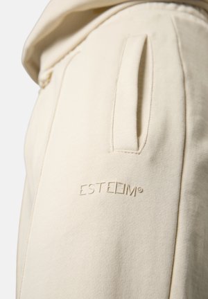 Cremefarbene Baumwoll-Jogginghosen mit einer seitlichen Tasche, geprägtem Logo "ESTEEM" am Oberschenkel und einer weichen, strukturierten Oberfläche.