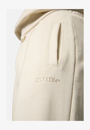 Cremefarbene Baumwoll-Jogginghosen mit einer seitlichen Tasche, geprägtem Logo "ESTEEM" am Oberschenkel und einer weichen, strukturierten Oberfläche.