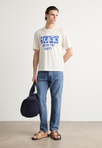 Beige bomull t-skjorte med blå grafisk tekst, kombinert med lyseblå jeans, brune sandaler og en rund marineblå duffelbag. Enkelt design.