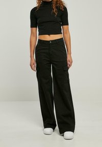 Schwarzes Crop-Top mit kurzen Ärmeln, kombiniert mit weiten schwarzen Cargo-Hosen mit Seitentaschen und weißen Sneakers. Glatte Stofftextur.