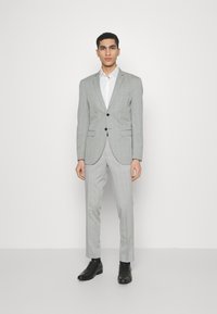 Pilkas languotas švarkas su derančiomis kelnėmis, balta marškinėliai su mygtukais ir juodi elegantiški batai. Lygi medžiaga, pritaikyta prie figūros, šiuolaikinis raštas.