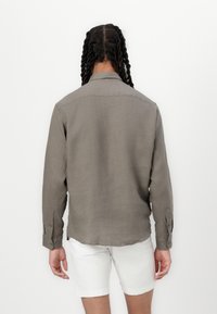 Camisa de lino gris con mangas largas, puños de botón y un collar, vista desde atrás. Llevada con pantalones cortos blancos.
