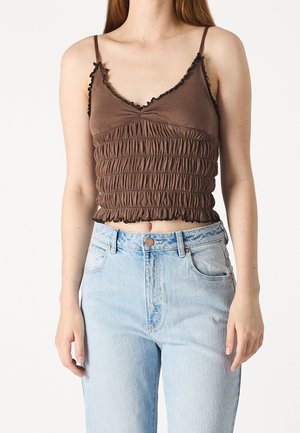 Brauner Crop-Top aus elastischem, gerafftem Stoff, mit dünnen verstellbaren Trägern und Rüschenkanten. Kombiniert mit hochgeschnittenen, hellblauen Jeans.