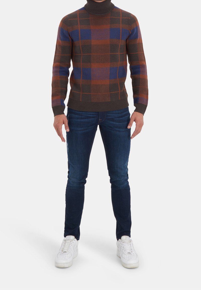 Hnědý a modrý kostkovaný svetr s rolákem a žebrovaným lemem, kombinovaný s tmavě modrými slim-fit džíny a bílými teniskami.