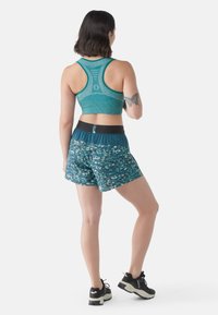 Sujetador deportivo en color azul verdoso y pantalones cortos estampados en azul verdoso con una cinturilla negra. La tela parece transpirable y tiene una textura suave. Zapatillas deportivas negras.
