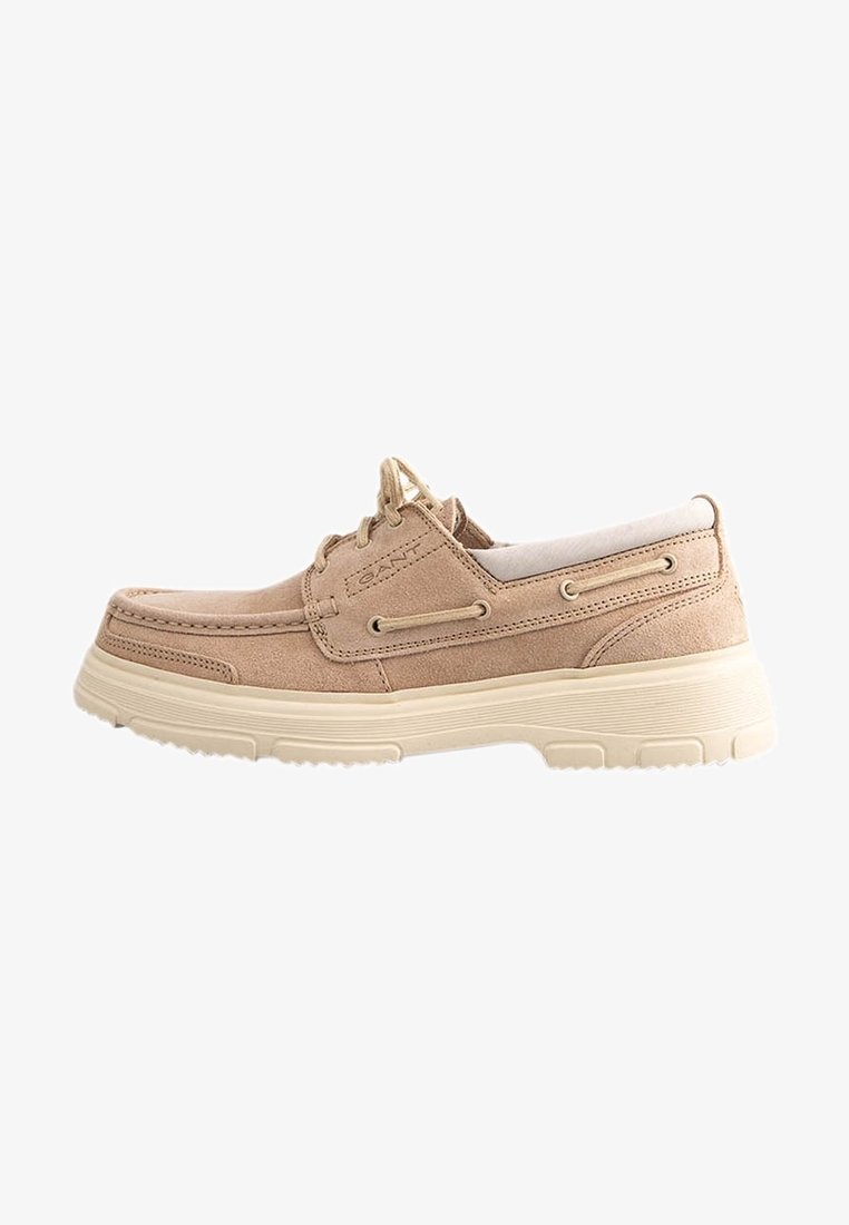 Chaussure slip-on en suède beige clair avec une tige texturée, une semelle blanche contrastante et des détails de laçage décoratifs sur les côtés.