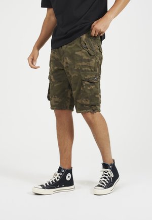 Tarnfarbene Cargo-Shorts in Dunkelgrün mit zwei Seitentaschen, einer Gesäßtasche und Knopfverschlüssen. Getragen mit schwarzen hochgeschnittenen Sneakers.