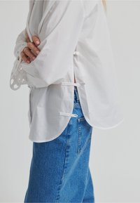 Chemise blanche en coton avec un ourlet arrondi, des fentes latérales et des détails décoratifs en corde blanche. Associée à un jean en denim bleu.