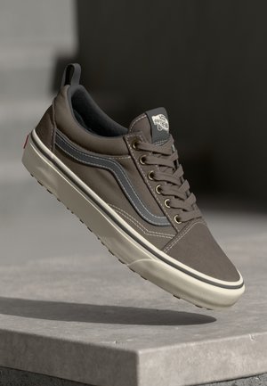 Brown Vans sneaker with white sole and gray stripe, mid-air above a concrete surface, highlighting the "Off The Wall" logo on the tongue.