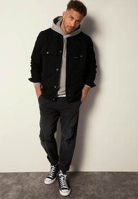 Zwarte lange mouwen jas over een grijze hoodie, gecombineerd met versleten zwarte broek en zwart-witte hoge sneakers. Eenvoudige achtergrond.