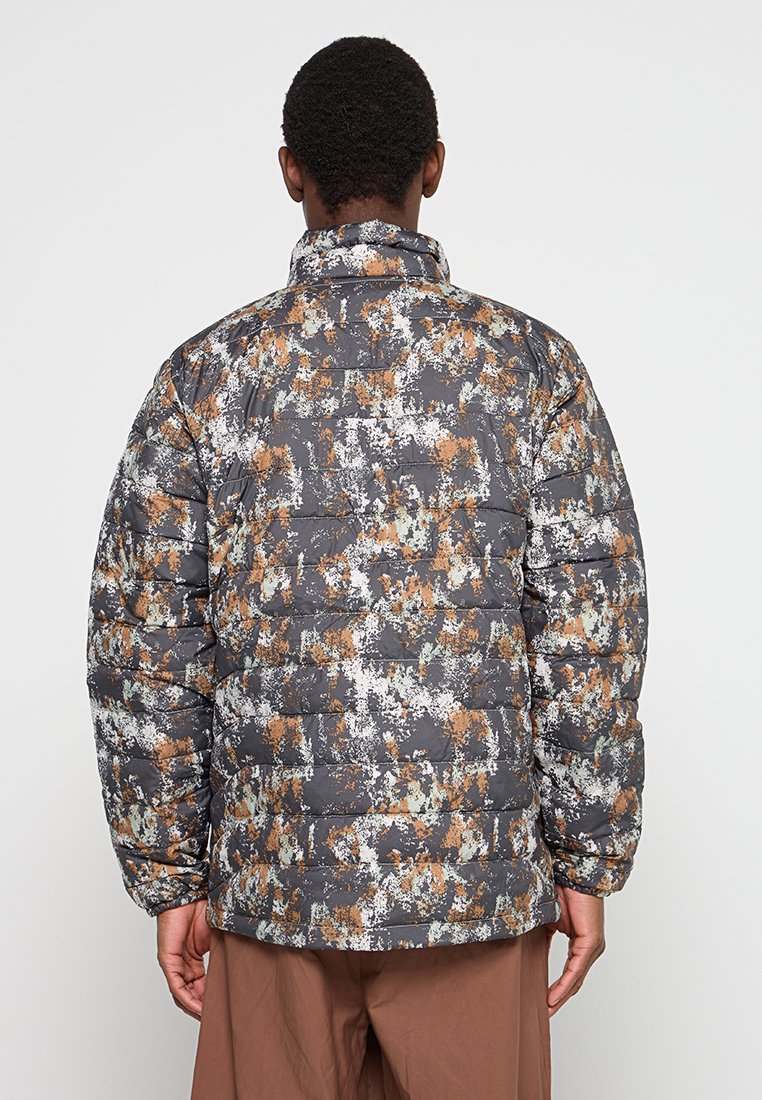 Osoba ubrana w wielokolorową kurtkę puchową w kamuflażu i brązowe spodnie, widoczna z tyłu na jednolitym tle.