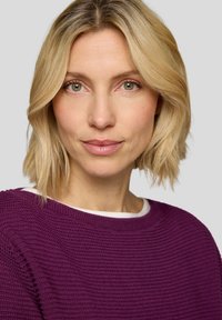 Lila Pullover mit strukturiertem Strick, rundem Ausschnitt und weißem Innenfutter. Kurze, wellige blonde Haare. Neutraler Hintergrund.