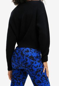 Schwarzer gestrickter Pullover mit weitem Schnitt und breiten Ärmeln, kombiniert mit blauen Hosen mit einem schwarzen Blumenmuster. Texturierte Stoffdetails sichtbar.