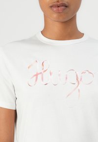 White cotton t-shirt featuring a pink ribbon-like text "Hugo" across the chest. Classic crew neckline and short sleeves. Smooth texture.