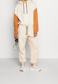 Felpa beige chiaro con maniche in sherpa arancione e logo; pantaloni jogger beige abbinati con strisce laterali arancioni; scarpe da ginnastica sportive; tiene una bottiglia d'acqua.